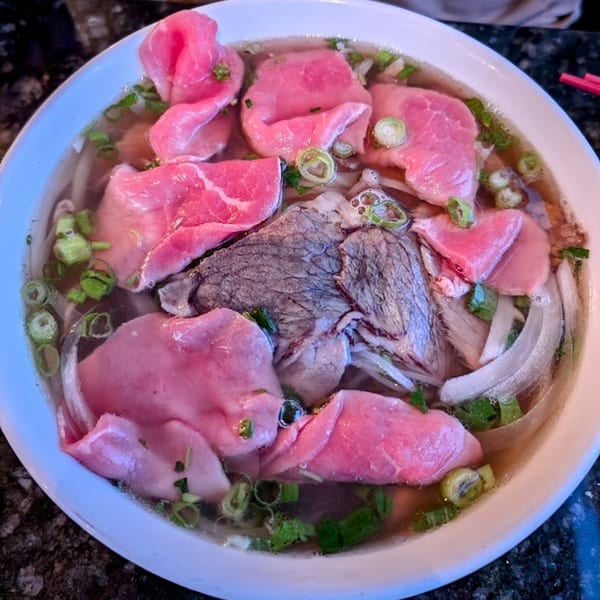 A close-up of a bowl of Vietnamese pho with rare beef slices, broth, noodles, and green onions.