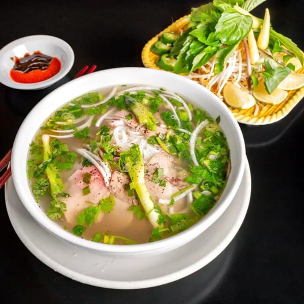A bowl of Pho with meat, noodles, and green onions, served with a side of fresh herbs.