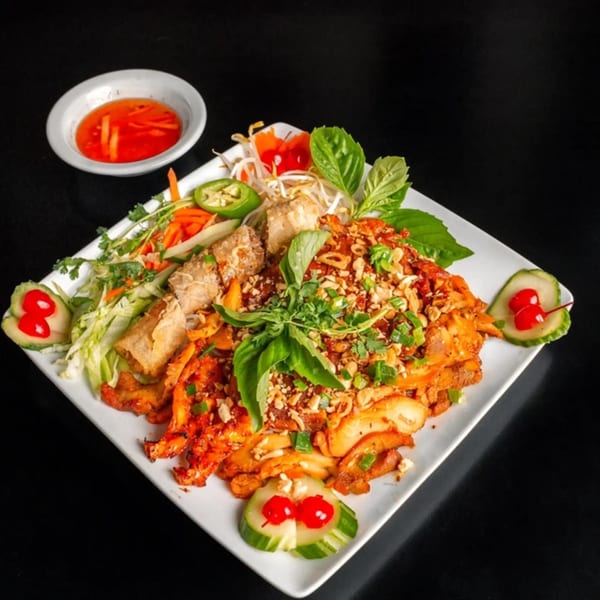 Vietnamese dish with chicken, spring rolls, vegetables, and a side of red dipping sauce.