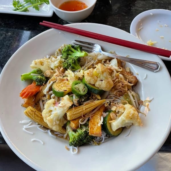 A Vietnamese noodle dish with broccoli, cauliflower, baby corn, zucchini, and tofu, served with red chopsticks.